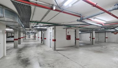 Beton duvarlar ve sütunlarda havalandırma kanalları, borular ve kırmızı güvenlik işaretleri olan temiz ve ferah yeraltı otoparkı.