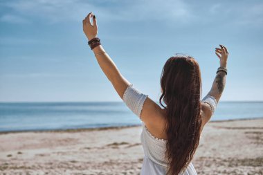 Sahilde sakin mavi okyanusla yüzleşirken genç bir kadının arka görüntüsü sakin bir farkındalık jesti olarak ellerini kaldırıyor. Özgürlük, okyanus tarafından ruhsal yenilenme