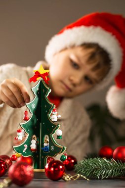 Noel Baba şapkalı çocuk ahşap bir yılbaşı ağacını oyuncaklarla süslüyor.