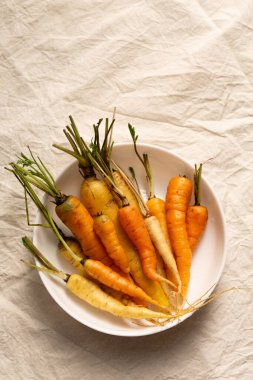 Kökleri ve sapları beyaz bir kâsede sıralanmış doğal turuncu ve sarı havuçlar orijinalliği ve tazeliği vurgular. Yavaş yaşam ve dikkatli bitki yeme kavramları için harikadır.
