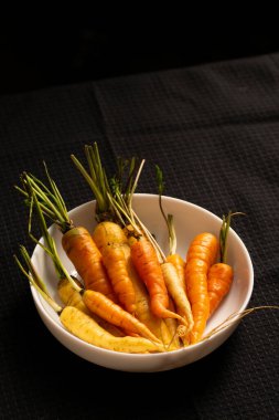 Taze toplanmış turuncu ve sarı havuçlar ve beyaz bir kasede sıralanmış yapraklı kapaklar dramatik bir kırsal yaşam yaratıyor. Yavaş yaşamak için mükemmel, çiftlikten sofraya ve dikkatli yemek kavramları için.