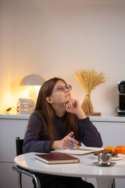 Genç bir öğrencinin beyaz bir masada oturup defterine düşüncelerini yansıttığı ve yazdığı sakin bir yaşam tarzı anı. Eğitim için mükemmel konsept, dikkatli planlama, yavaş yaşam, zihinsel sağlık