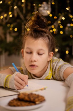 Sıcak süveter giyen konsantre genç kız Noel Baba 'ya Noel ağacının yanında Noel mektubu yazıyor.