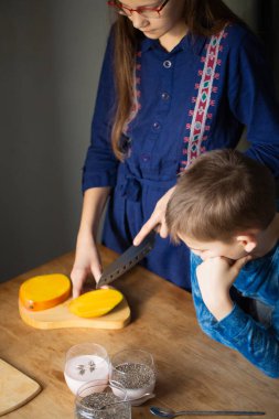 Küçük kardeşi izlemek için eğilirken, tahta bir tahtanın üzerinde olgun mangoyu dikkatlice kesen kız, ev yapımı sağlıklı bir tatlı hazırlamak için bir bardak bitki sütü ve chia tohumuyla hazır bekliyor.