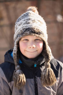 Kış şapkalı gülümseyen bir çocuğun yakın plan portresi karla kaplı, çocukluk sevincini, sıcaklığı ve mutlu kış anılarını simgeliyor..