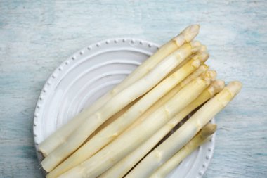 Beyaz seramik tabakta taze beyaz kuşkonmaz, açık mavi ahşap arka plan, mevsimsel organik üretim kavramı.