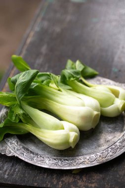 Kırsal ahşap masa üzerindeki dekoratif metal tabakta taze yeşil bok choy, doğal ışık, organik mevsimlik sebze.