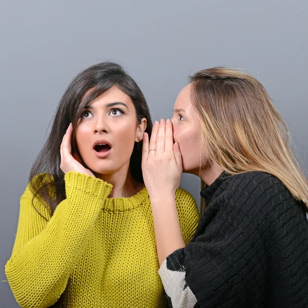 Дві жінки друзі шепочуть секрети на сірому фоні — стокове фото