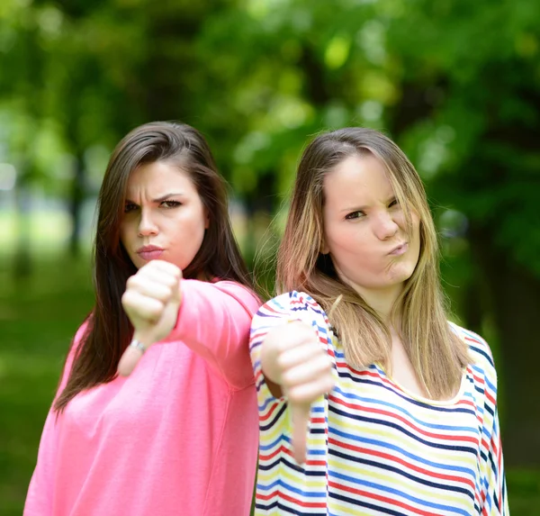 Due ragazze attraenti mostrano pollici verso il basso contro la natura verde backg — Foto Stock