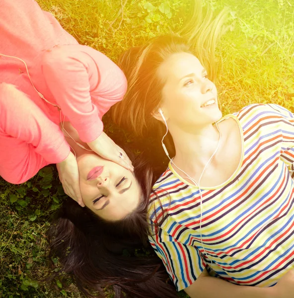 Two girl friends listening to music while lying on grass during — Stock Photo, Image