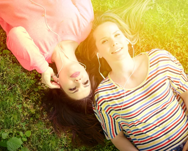 Two girl friends listening to music while lying on grass during — Stock Photo, Image