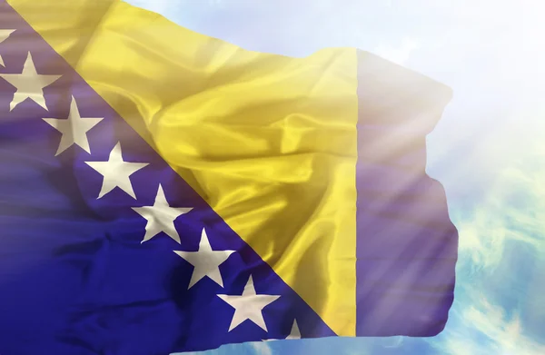 Bosna a Hercegovina mává vlajkou proti modré obloze se slunečními paprsky — Stock fotografie