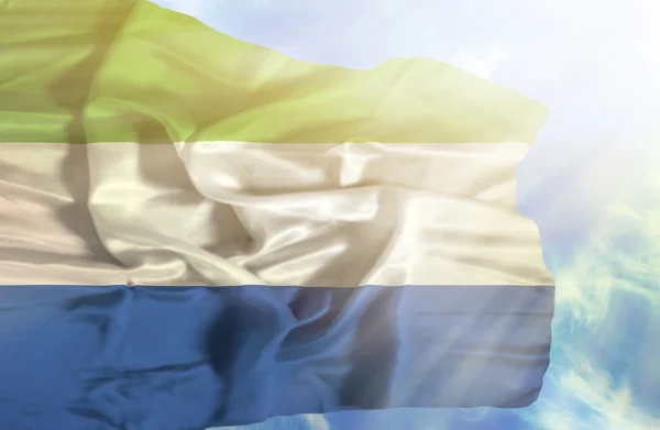 Sierra Leone schwenkt Flagge gegen blauen Himmel mit Sonnenstrahlen — Stockfoto