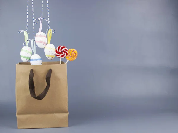 Home decor for Easter. Shoping bag with bright Easter eggs hanging on strings on a gray background, copy space