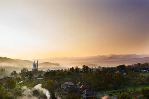 Aerial view of village in fog with golden sunbeams at sunrise in autumn. Beautiful rural landscape with foggy colorful trees, church, orange sky with sun. Fall in Romania.