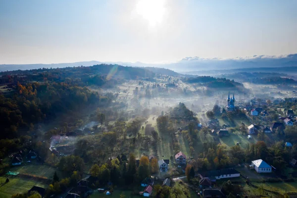 秋の日の出に黄金の太陽の光で霧の中の村の空中ビュー 霧のようなカラフルな木々 太陽とオレンジの空と美しい田園風景 ルーマニアの秋 — ストック写真