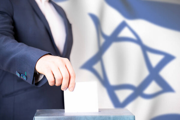 Election in Israel. Hand of voter putting vote in the ballot box. Waved Israel flag on background.