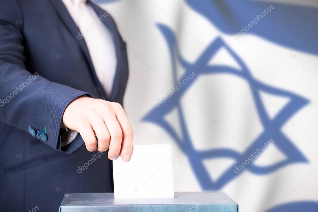Election in Israel. Hand of voter putting vote in the ballot box. Waved Israel flag on background.