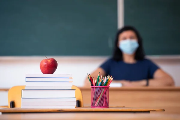 Back to School Concept. Apple, accessories and books in the classroom, teacher wearing mask in the background.