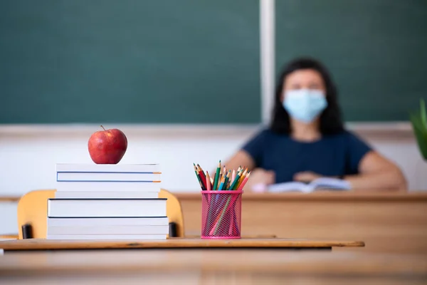 Back to School Concept. Apple, accessories and books in the classroom, teacher wearing mask in the background.