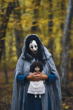 Cadılar Bayramı konsepti: Korkunç maskeli ve garip elbiseli, ürkütücü bir bebek tutan, sonbahar ormanında duran hayalet kadın..