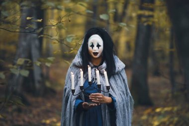 Cadılar Bayramı konsepti, korkunç maskeli, garip elbiseli, elinde mumlarla sonbahar ormanlarında dikilen hayalet bir kadın..