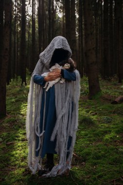 Cadılar Bayramı konsepti: Korkunç maskeli ve garip elbiseli, ürkütücü bir bebek tutan, sonbahar ormanında duran hayalet kadın..