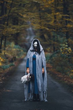 Cadılar Bayramı konsepti: Korkunç maskeli ve garip elbiseli, ürkütücü bir bebek tutan, sonbahar ormanında duran hayalet kadın..