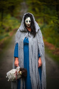 Cadılar Bayramı konsepti: Korkunç maskeli ve garip elbiseli, ürkütücü bir bebek tutan, sonbahar ormanında duran hayalet kadın..