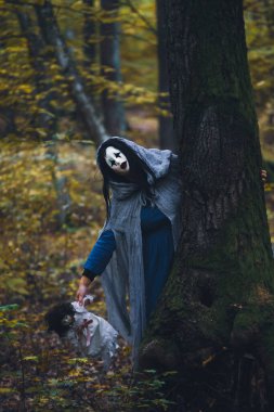 Cadılar Bayramı konsepti: Korkunç maskeli ve garip elbiseli, ürkütücü bir bebek tutan, sonbahar ormanında duran hayalet kadın..