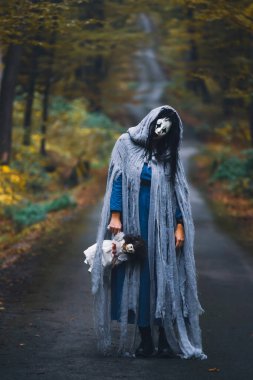 Cadılar Bayramı konsepti: Korkunç maskeli ve garip elbiseli, ürkütücü bir bebek tutan, sonbahar ormanında duran hayalet kadın..