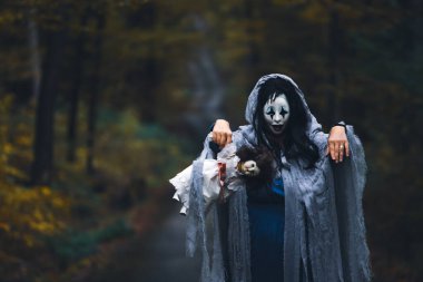 Cadılar Bayramı konsepti: Korkunç maskeli ve garip elbiseli, ürkütücü bir bebek tutan, sonbahar ormanında duran hayalet kadın..