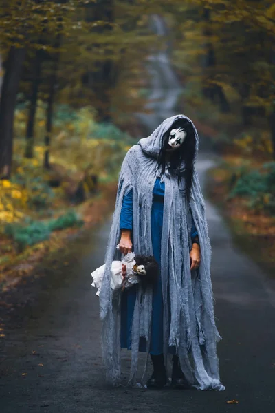Cadılar Bayramı konsepti: Korkunç maskeli ve garip elbiseli, ürkütücü bir bebek tutan, sonbahar ormanında duran hayalet kadın..