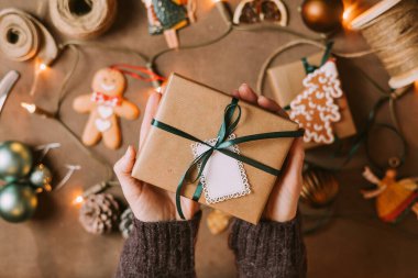 Noel hediyesi kutusuyla çevrili, sıcak atmosferde ışıldayan Noel ışıkları ve şenlikli dekorasyonlarla çevrili kadın elleri, Noel arifesi hazırlıkları.