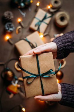 Noel hediyesi kutusuyla çevrili, sıcak atmosferde ışıldayan Noel ışıkları ve şenlikli dekorasyonlarla çevrili kadın elleri, Noel arifesi hazırlıkları.