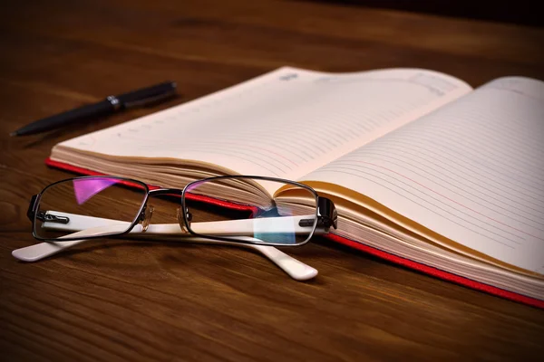 Diario rojo abierto, gafas y bolígrafo en la mesa de madera, de cerca —  Fotos de Stock