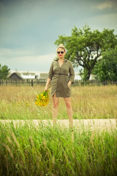 Una joven con un vestido verde de pie con un ramo de flores amarillas en la mano. Vida de pueblo en el campo . —  Fotos de Stock