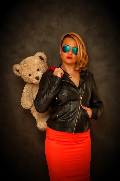 Glamour beautiful girl-blonde in a leather jacket, sunglasses and a red skirt with a teddy bear on a dark background — Stock Photo, Image