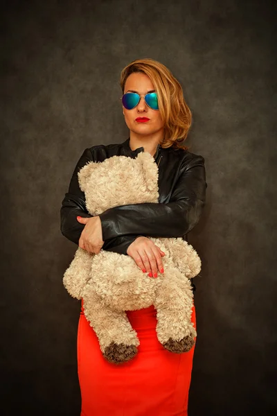 Chica rubia de moda en una chaqueta de cuero, gafas de sol y una falda roja con un oso de peluche en sus brazos —  Fotos de Stock