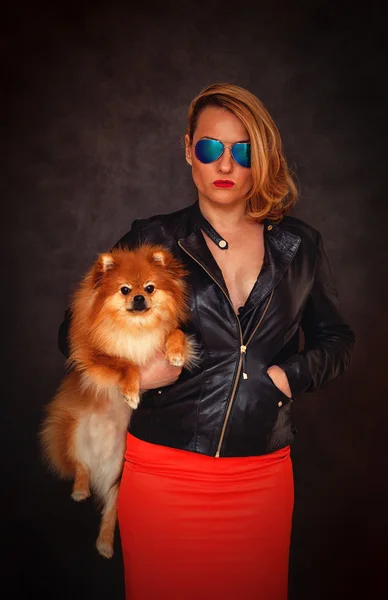 革のジャケット、サングラス、赤いスカートに厳格な魅力的な女性が彼女の腕の中で犬を保持します。 — ストック写真