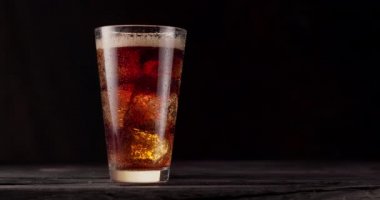 Buzlu bir bardak kola siyah arka planlı ahşap bir masanın üzerinde duruyor. Gaz kabarcıkları bardakta aktif olarak yükselir. Ağır çekim, Blackmagic Ursa Pro G2.