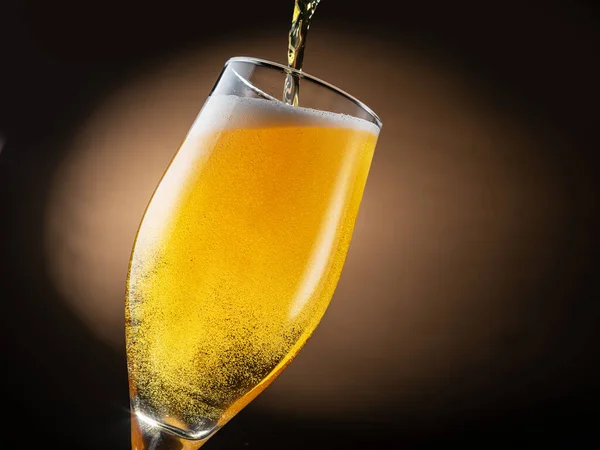 Jet of beer is poured into a beer glass, causing a lot of bubbles and foam. Dark brown background.