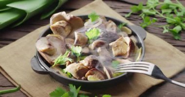 Kızarmış porcini mantarlı tava eski bir ahşap masada duruyor ve buhar yayıyor. Bir adamın eli çatalla bir mantar topluyor.. 