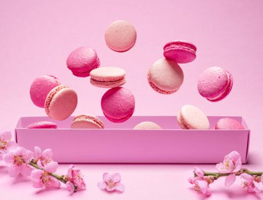 Renkli tatlı badem kurabiyeleri, karton kutunun üstünde havada uçuşan lezzetli kurabiyeler. Kutunun yanında çiçek açan kiraz dalı.