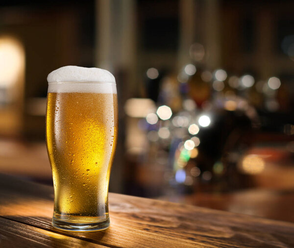 Cooled glass of pale beer with condensation drops on glass surface on the wooden table. Blurred bar at the background.