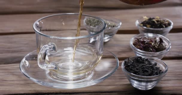 急須からゆっくりとグラスカップに茶を注ぎ 周りにはさまざまな種類の茶葉を入れた茶碗 ヴィンテージの木製テーブルがあります — ストック動画
