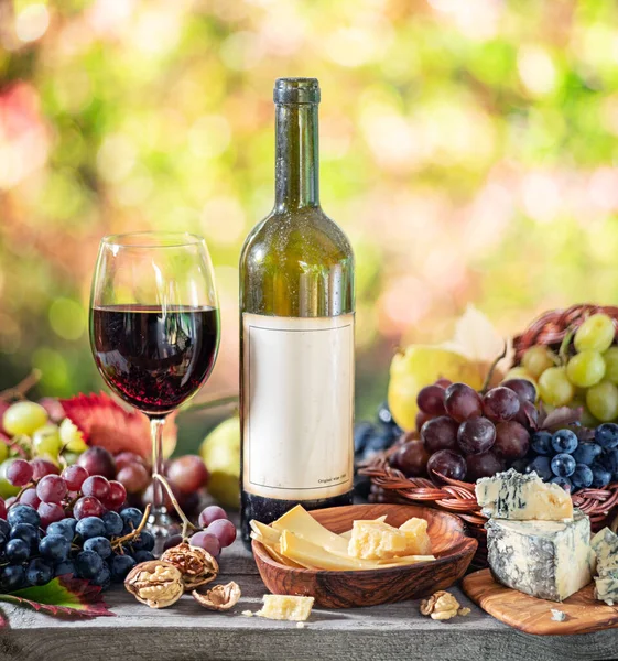 Grapes, bottle of wine and different cheeses on country wooden table and blurred colorful autumn background. Variety of products as the symbol of autumn abundance and prosperity.