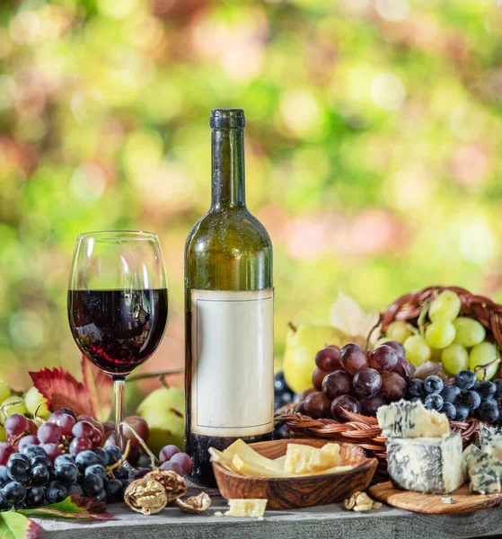 Grapes, bottle of wine and different cheeses on country wooden table and blurred colorful autumn background. Variety of products as the symbol of autumn abundance and prosperity.