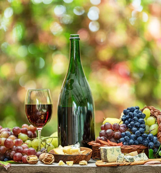 Grapes, bottle of wine and different cheeses on country wooden table and blurred colorful autumn background. Variety of products as the symbol of autumn abundance and prosperity.