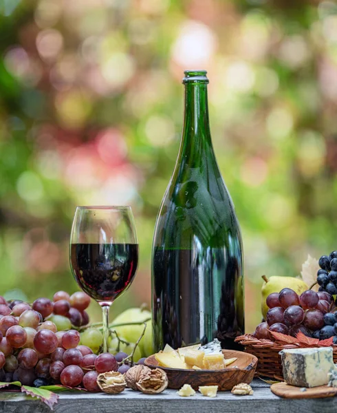 Grapes, bottle of wine and different cheeses on country wooden table and blurred colorful autumn background. Variety of products as the symbol of autumn abundance and prosperity.